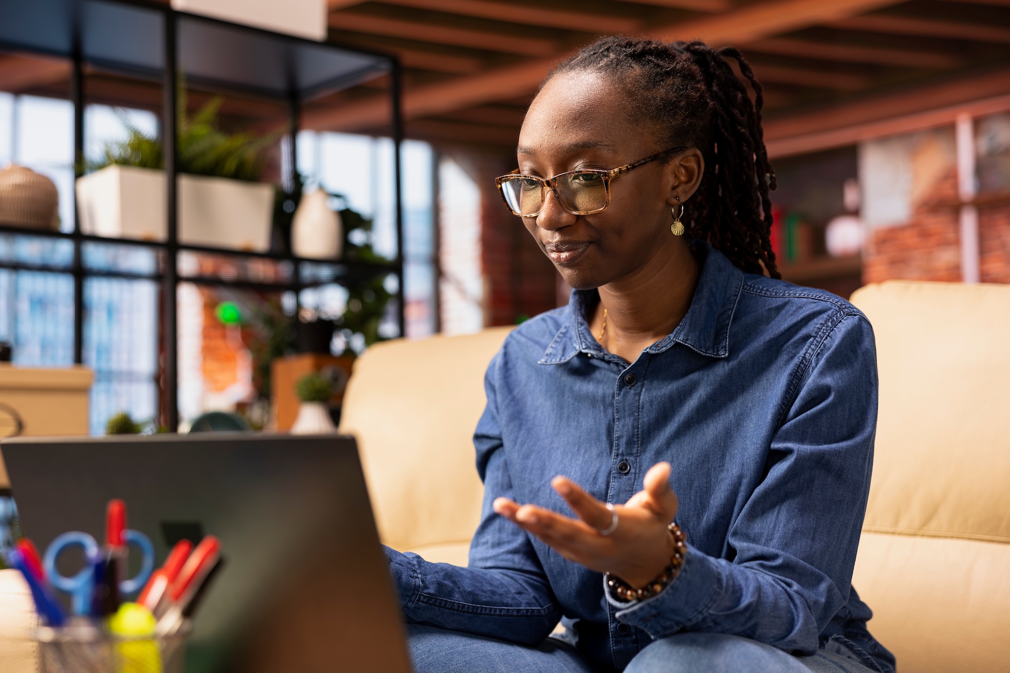 A woman wearing a blue denim shirt gesturing while participating in a video call on her laptop.